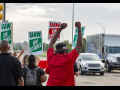 Strikers shown from behind, waving at cars, fists in air, signs read "UAW On Strike"