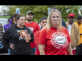 A group of autoworkers stand around outside looking at the camera--the closest is a blonde woman with a red shirt that says “UAW Back in the Fight.”