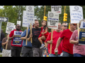 A group of people in red shirts, one man carrying a child march past the camera with signs saying Stellantis is barganing in bad faith.