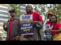 A group of workers march in Detroit holding signs that say "UAW Stand Up: Fair Pay and COLA Now." 