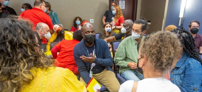 Masked people sit in small groups in a conference room, talking. They are diverse in apparent race, age, and gender.