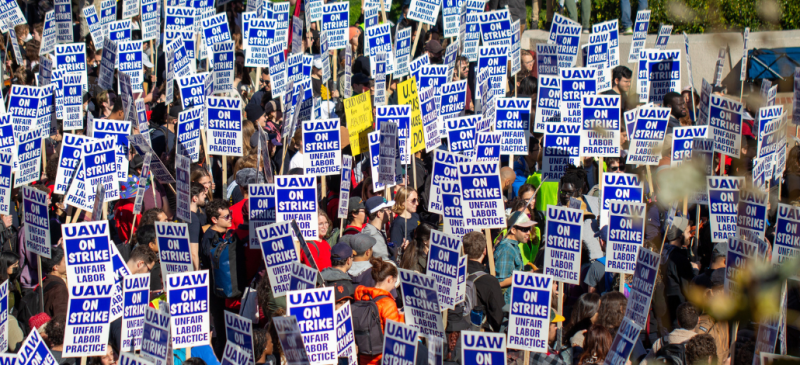 Marching crowd viewed from above, all holding printed picket signs that say "UAW on strike, unfair labor practice"
