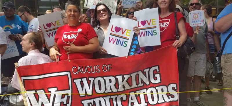 Three smiling people stand among a crowd outdoors. They hold a big red banner that says "Caucus of Working Educators" with a "WE" logo and two wear read shirts iwth the same logo. Two hold white signs with rainbow letters: "Love wins" with a heart for the letter O.  Signs carried by others behind them say "I [heart] my trans son," something ending in "LGBTQ and Trans People," and "Peace."