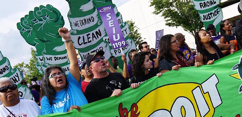 Crowd at rally behind bright green banner, woman with fist up, people holding signs, one SEIU sign visible, another for racial justice, picture at an angle