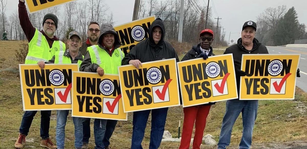 Seven workers, men and women, white and black, hold yellow “union yes” signs in front of the Lordstown water tower