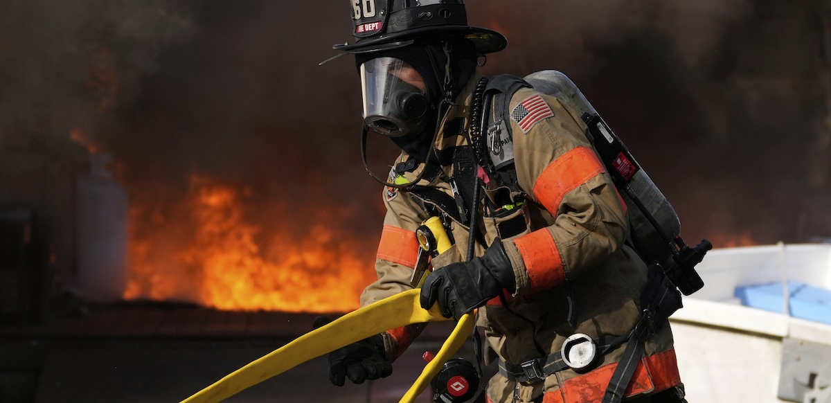 A firefighter in full protective equipment moves a yellow hose with fire in the background