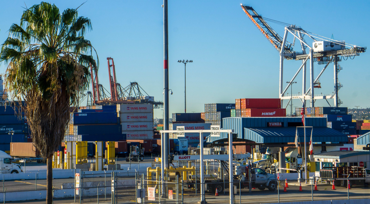 A photo of the ports of L.A. and Long Beach, with a big crane in the background and containers in the foreground, and a palm tree on the far left.