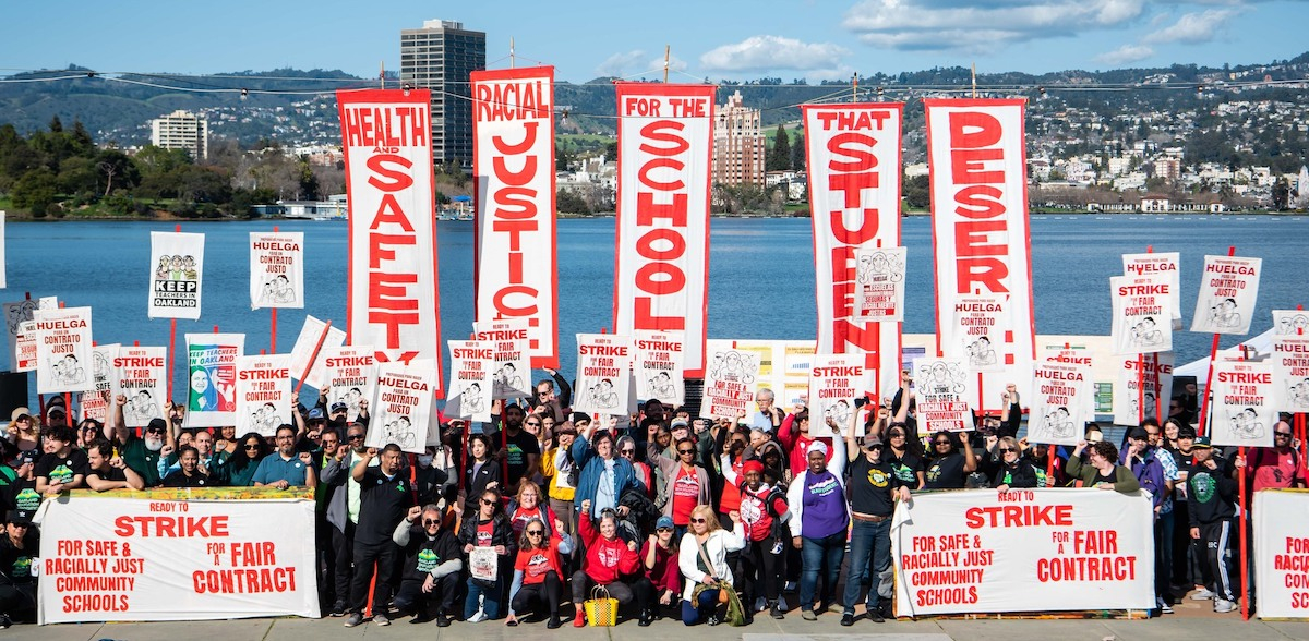 A crowd of hundreds is seen from afar and behind them there’s a body of water. They have vertical red on white banners saying “Health and Safety,” “Racial Justice,” “For the schools,” “That Students,” “Deserve.” 