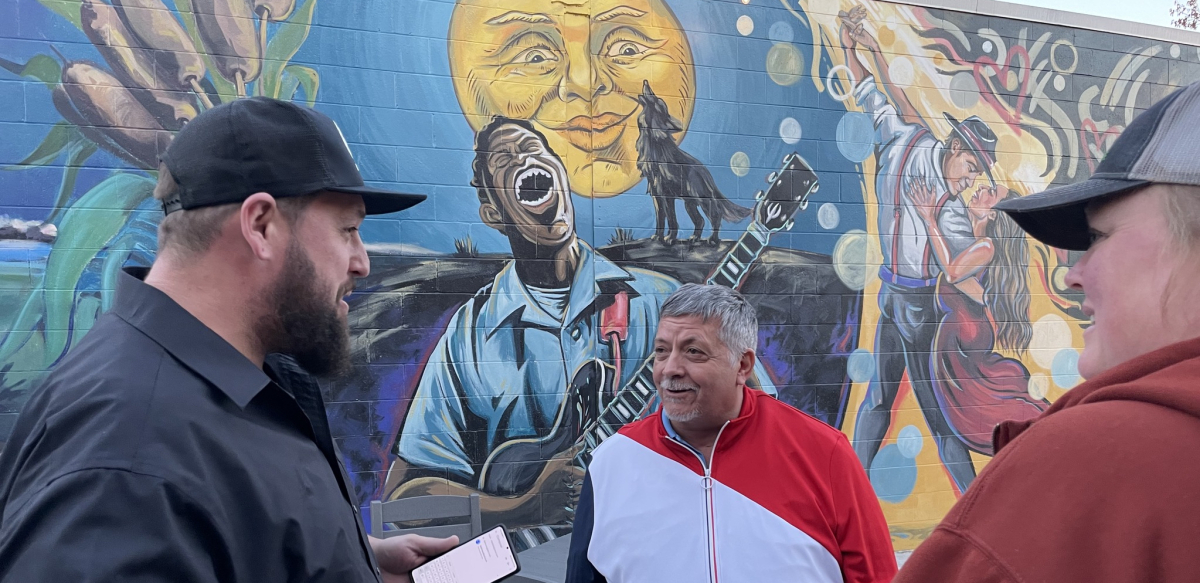 Three autoworkers talk while standing in front of a colorful mural with a painting of a man singing and playing guitar, a wolf howling, and a couple dancing the tango.