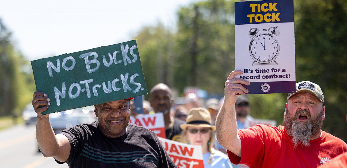 Auto workers with signs saying no bucks, no trucks and Tick Tock Time for a Record Contract march towards the camera