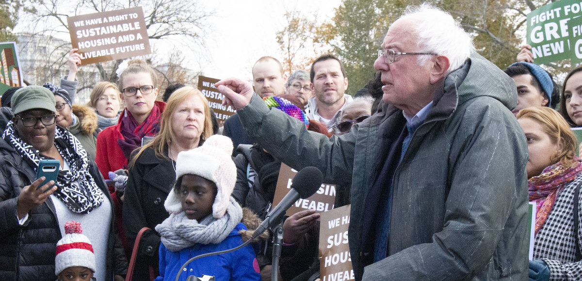 Bernie Sanders speaks at a podium surrounded by people holding signs about housing