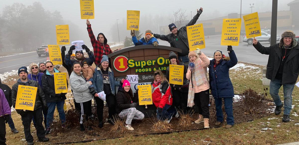 A group of 20 workers in winter clothes gather with yellow UFCW signs around a SuperOne Foods sign