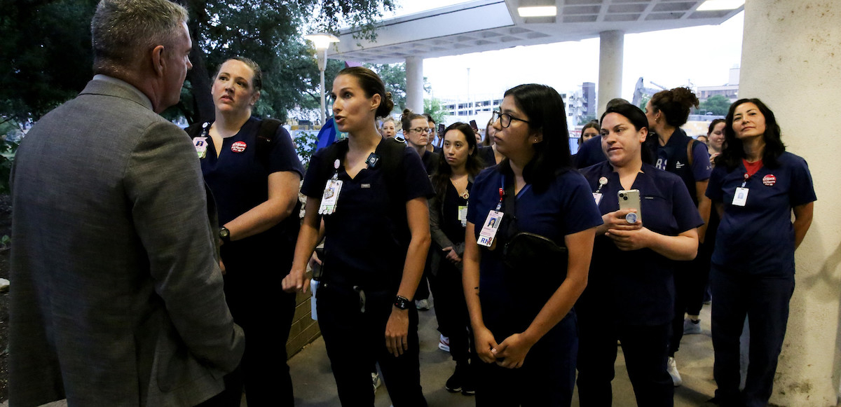 A group of nurses in dark blue scrubs with badges confront a man in a grey suit who is blocking their path.