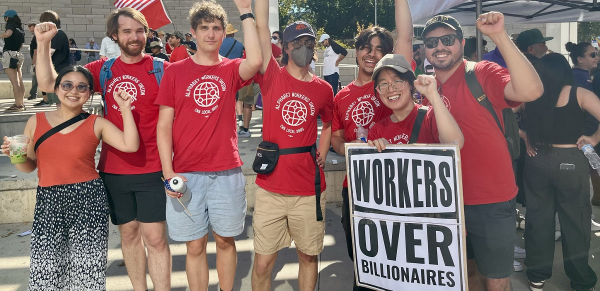 A group of Google workers stand in red shirts behind a sign that reads Workers Over Billionaires.