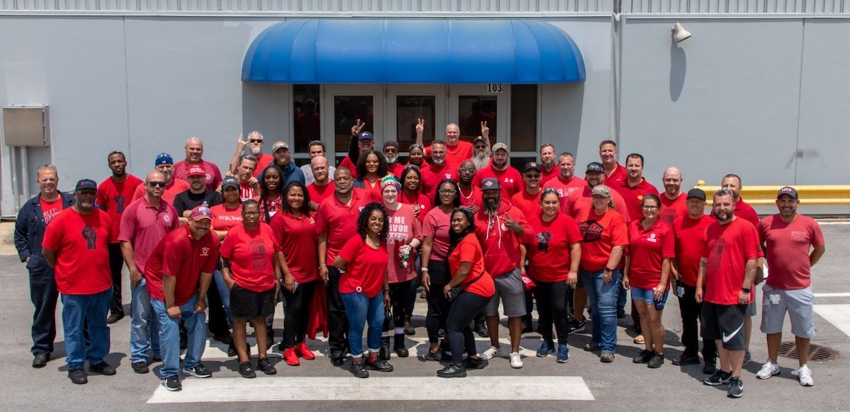 Forty workers in red t-shirts pose together outside a building with a blue awning.