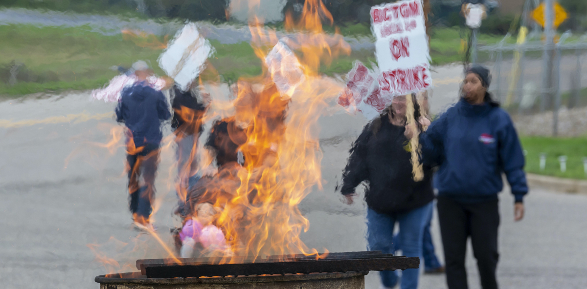 Workers holding picket signs that read BCTGM Local 3G On Strike march in the background, while a burn barrel with flames sits in the foreground.