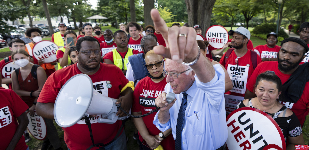 Bernie Sanders speaks into a megaphone, with his finger pointed towards the top of the frame, surrounded by red-shirted members of the union UNITE HERE.