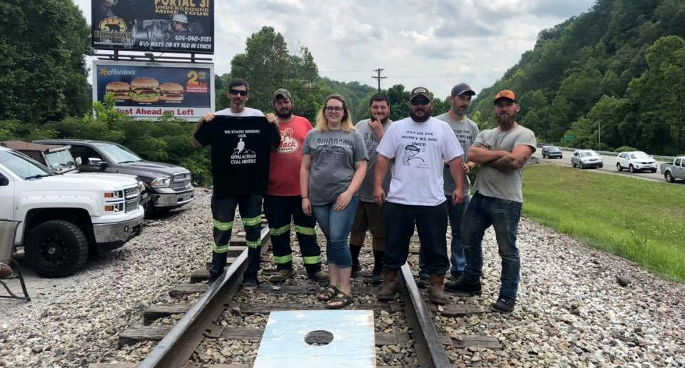 Five people stand on railroad tracks, with a cornhole game at their feet. Some wear "pay us what we are owed" shirts.