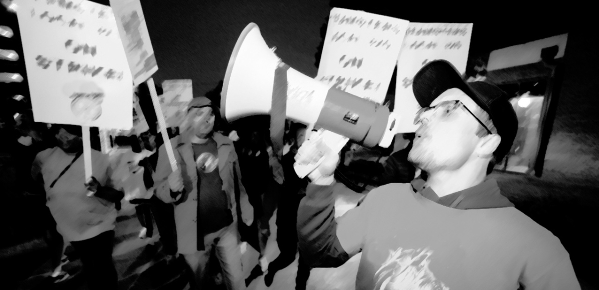 A man yells into a bullhorn with people in the background holding signs--is it blurry and black and white
