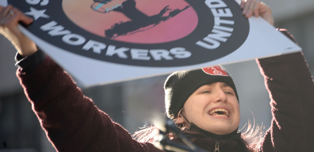 Woman in a warm hat holds Starbucks Workers United sign high with sun behind her