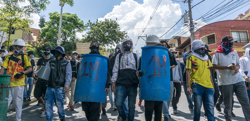 Row of protesters blockading the street in Colombia, two of whom are wielding blue shields of some kind.