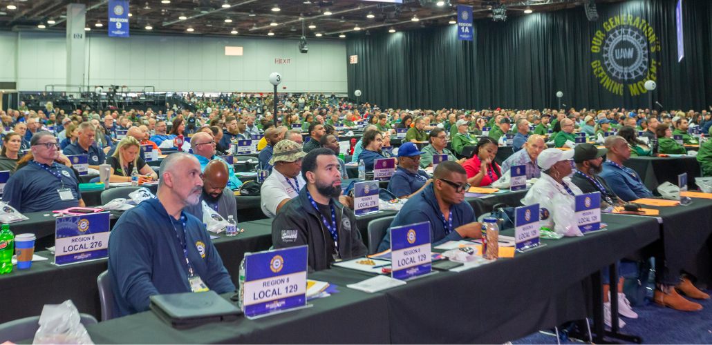 UAW delegates (men and women, Black and white) sit listening at the union's 2026 convention. Table signs identify those in the foreground as from Region 8, Locals 129 and 276.