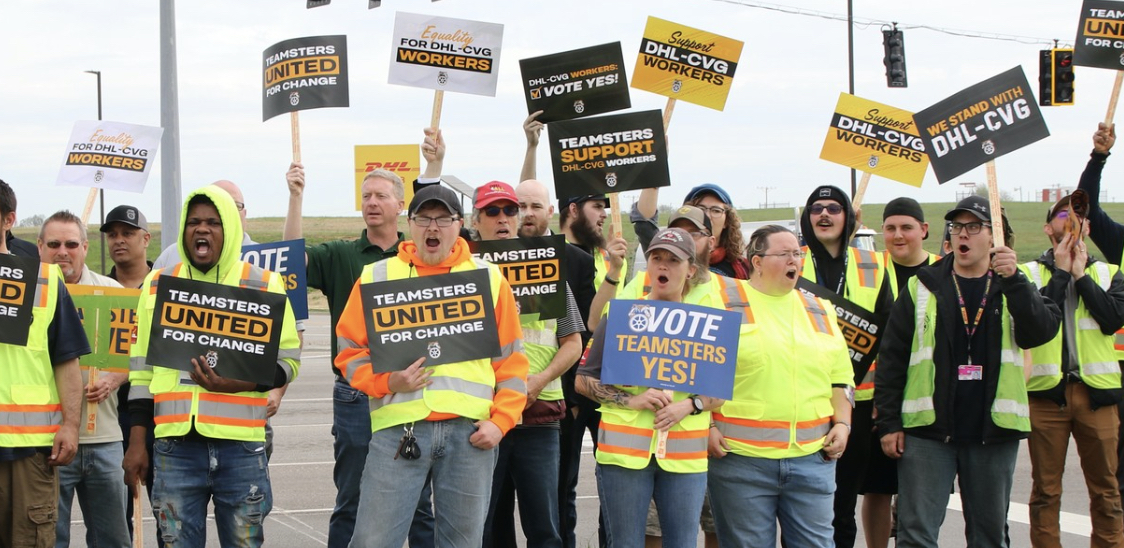 A group of 15 or so workers in hi-viz vests look excited and hold signs about voting for the Teamsters union.