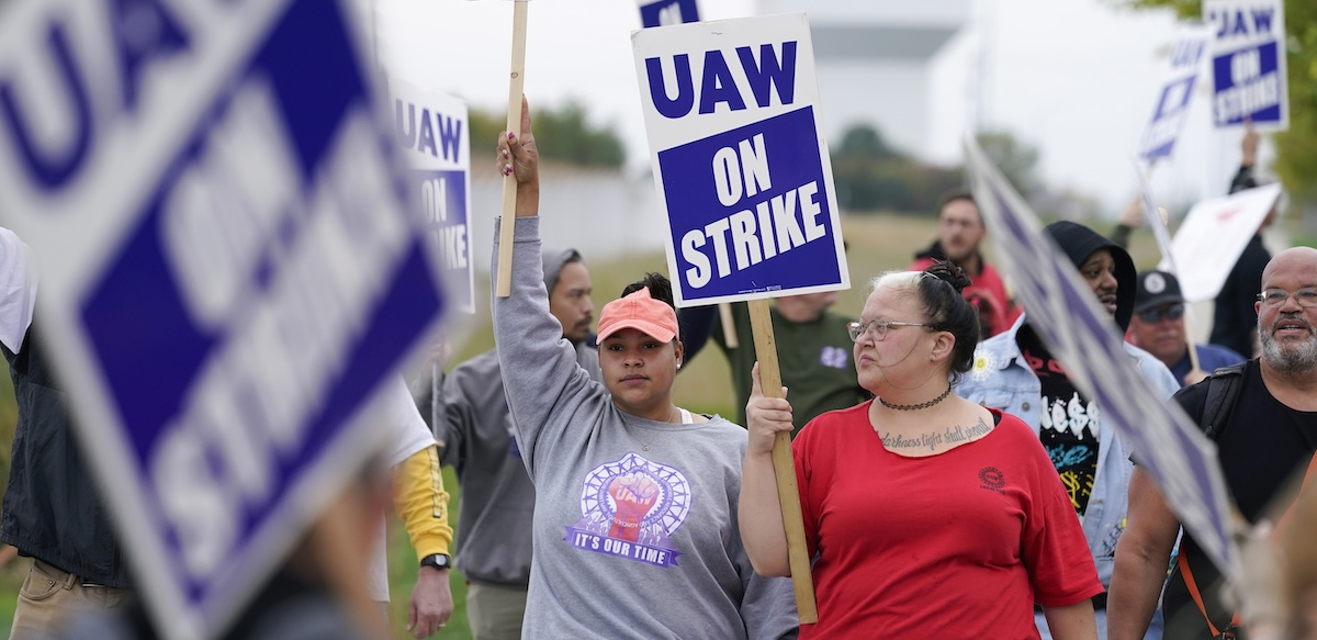 The camera focuses on two women in a crowd of picketers, diverse in race and gender, carrying printed blue and white "UAW on strike" picket signs. The women are wearing different UAW T-shirts. One has the collar cut off, revealing part of a tattoo that reads, in cursive, "darkness light shall prevail." Both women wear serious, determined expressions.