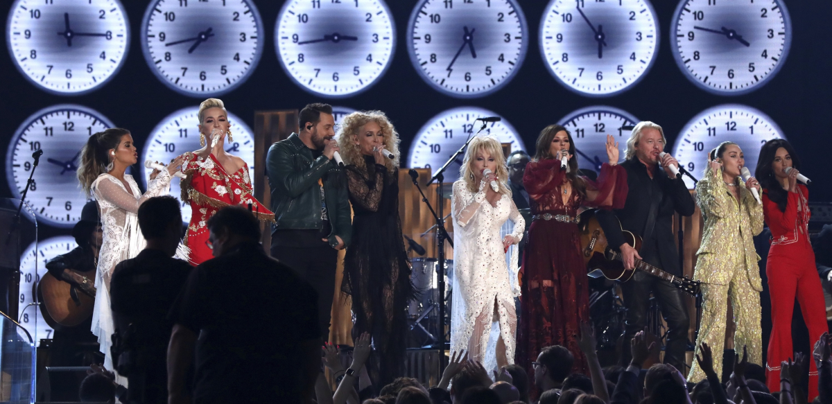 A row of musicians with large clocks behind them perform, with Dolly Parton in the center