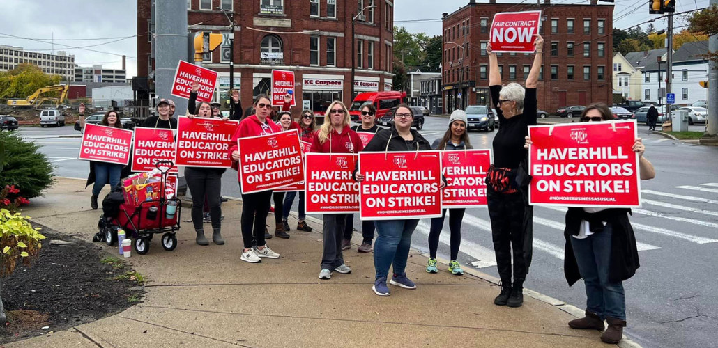 About 15 people, mostly white women, stand on a street corner wearing big red printed signs that say "Haverill educators on strike!" One woman holds her sign high in the air and turns to face the others, in profile to the camera, with a lively open-mouthed expression, maybe yelling or singing. Her sign says "Fair contract now." 
