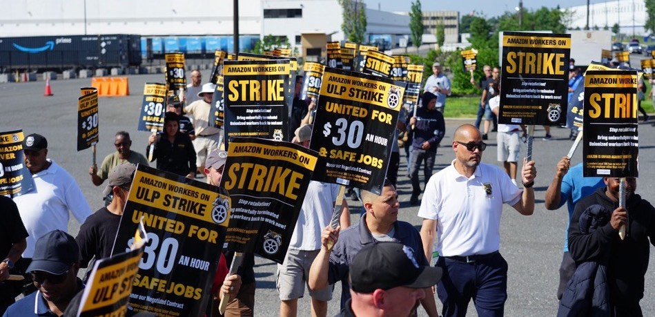 A big group of men marches, carrying black-and-yellow picket signs that say: "Amazon workers on ULP strike, united for $30 an hour and safe jobs" with a Teamsters logo