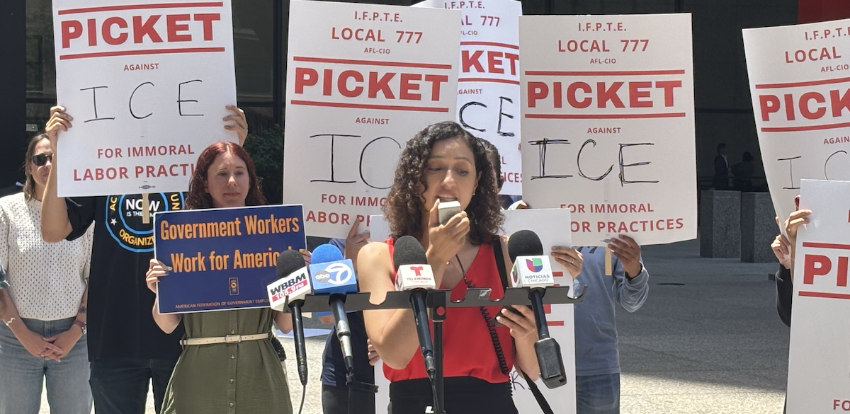 A woman speaks into a microphone outside while others stand behind her with signs that say “Picket ICE”