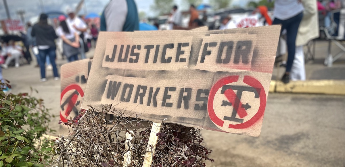 A hand-stenciled sign in the foreground says “Justice for Workers” while in the background striking workers stand outside a plant