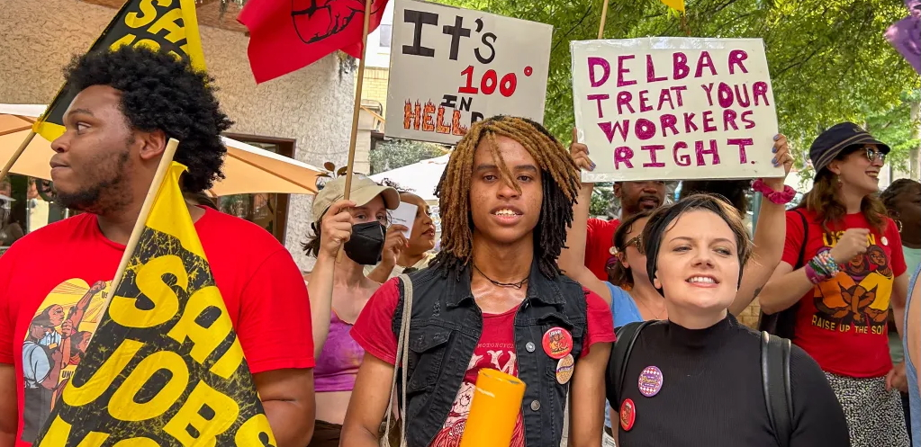 Several people with colorful signs in red and orange face the camera, one says “Delbar treat your workers right”