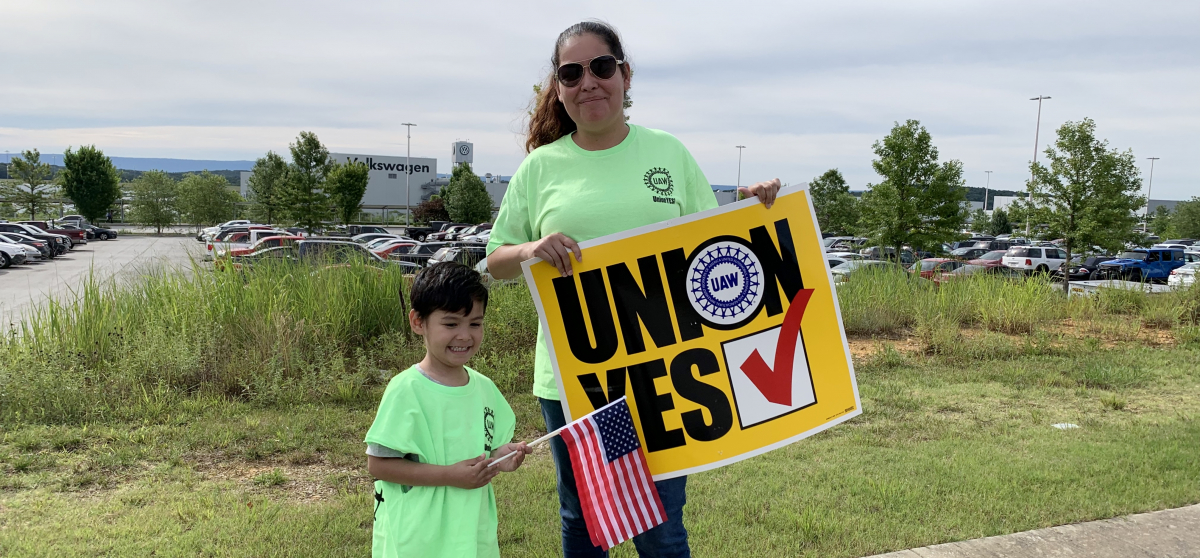 Woman holding "UNION YES" sign and child holding U.S. flag, with VW plant behind