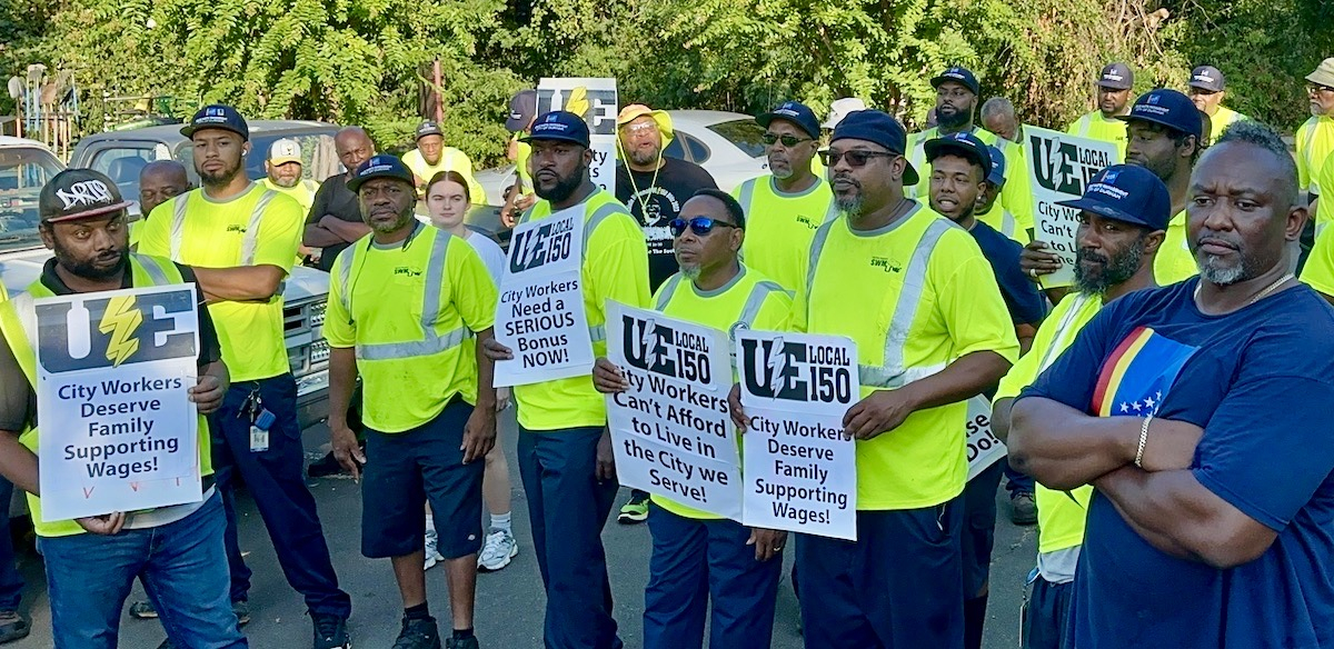 A group of sanitation workers, most in their bright green hi-visibility work vests, hold signs saying, "Fair Pay for City Workers!" and "City Workers Can't Afford to Live in the City we Serve!" during a rally.