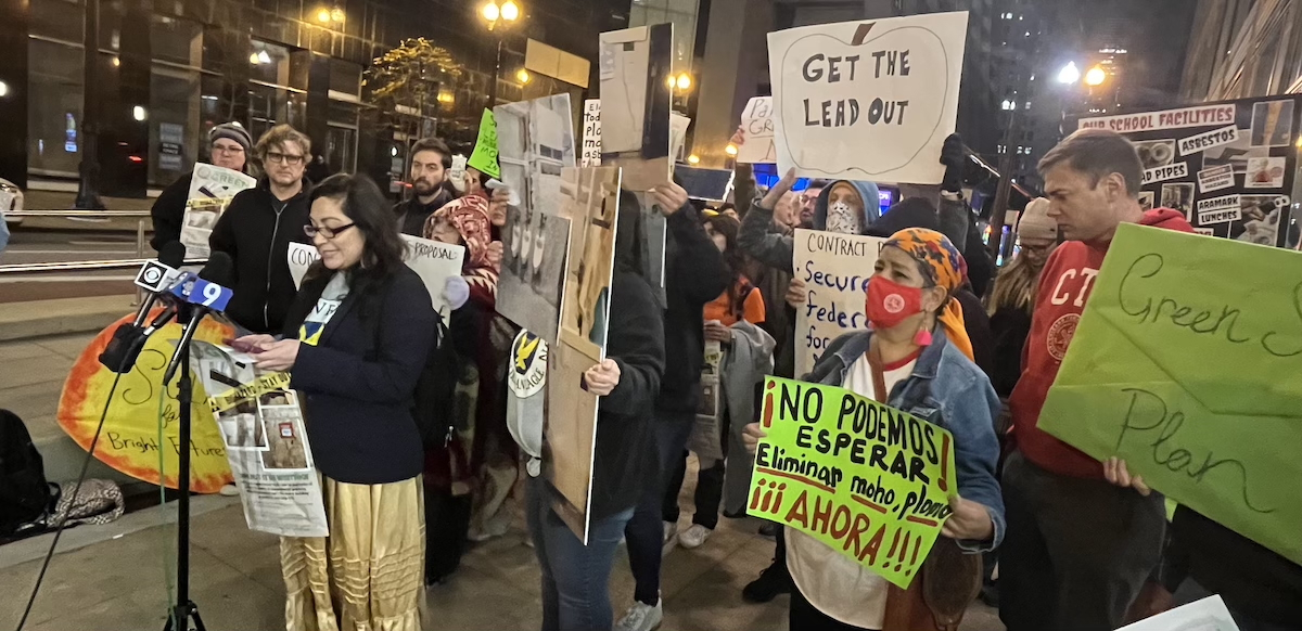 a speaker at a mic set up on the sidewalk is backed up by people holding signs about lead in schools