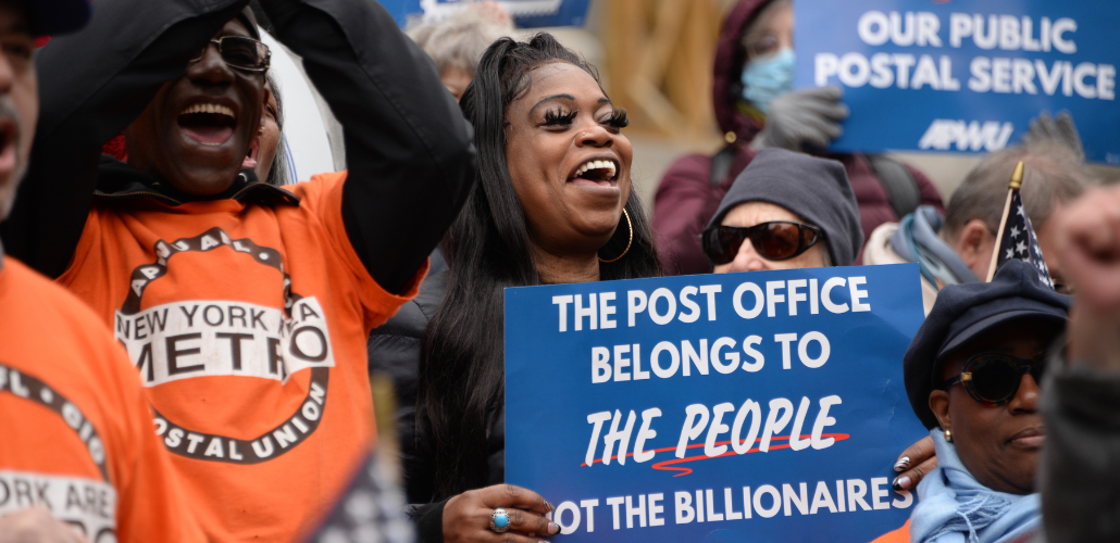 Two Black workers foregrounded in a dense crowd. Man on left wears orange New York Metro APWU T-shirt and lifts his hands to clap. Woman on right has thick eyelashes and holds a blue printed sign: "The Post Office belongs to the people, not the billionaires." Both smiling, energetic, probably chanting.