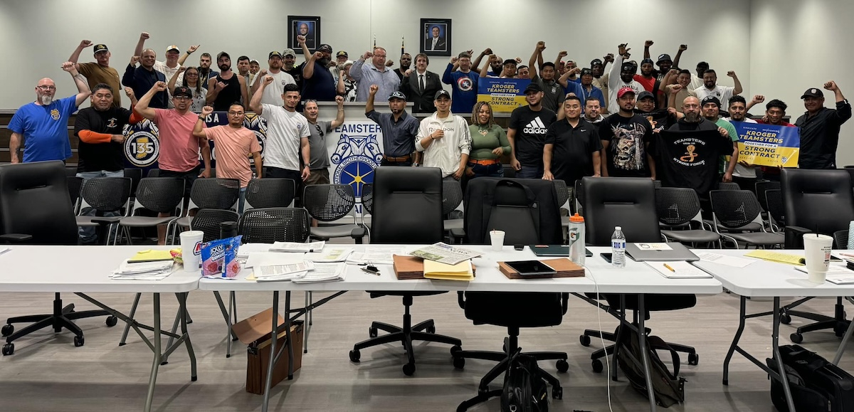 A group of 50 people stand behind a bargaining table with fists raised. 
