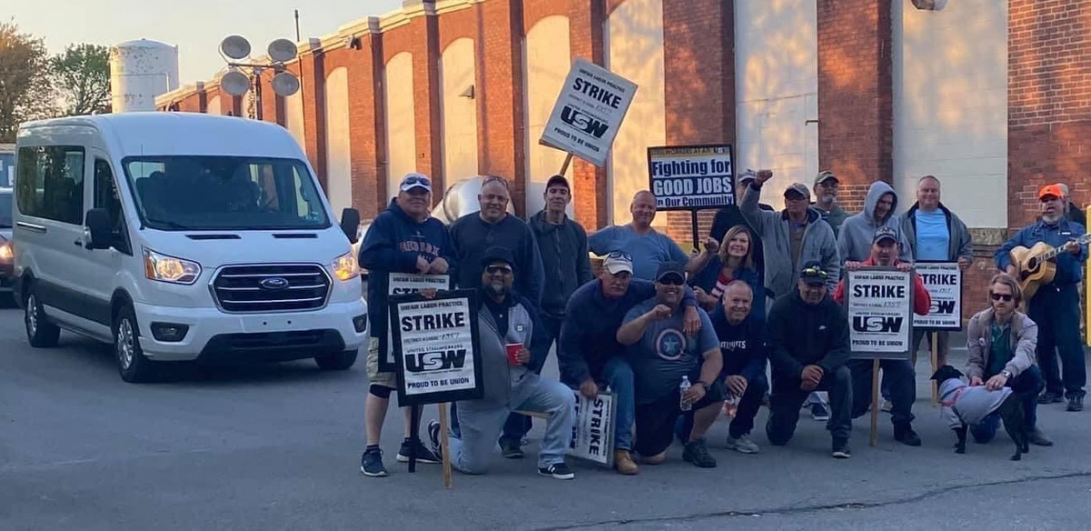 A group of striking steelworkers pose for a photo in a driveway, while a van carrying replacement workers waits behind them.