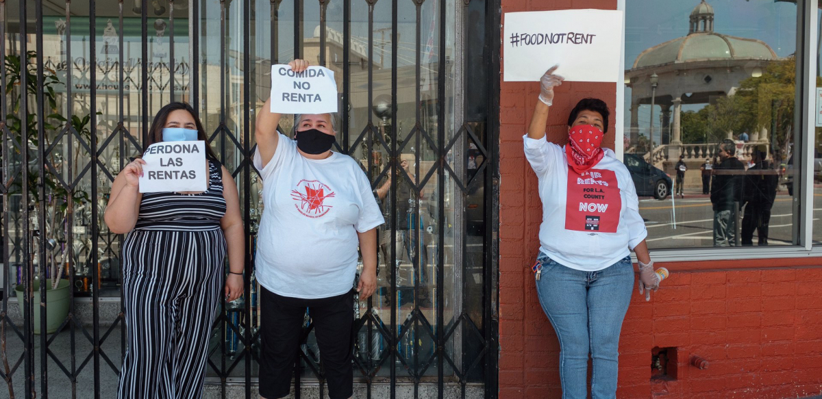 Three women hold signs saying "Food Not Rent" and "Cancel Rent" in English and Spanish.