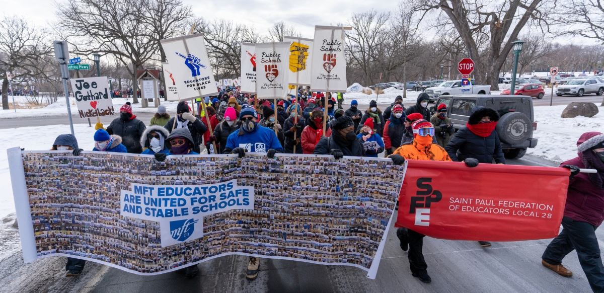 People march in the street on a snowy day. Those at the front carry two huge banners: MFT banner has blue union logo and hundreds of tiny images of members holding signs. SFPE one is red. Others in crowd are holding up beautiful hand-painted signs on sticks with messages like "Educators: the heart of the community" (with a fist/heart image) and "Educators for Black lives"