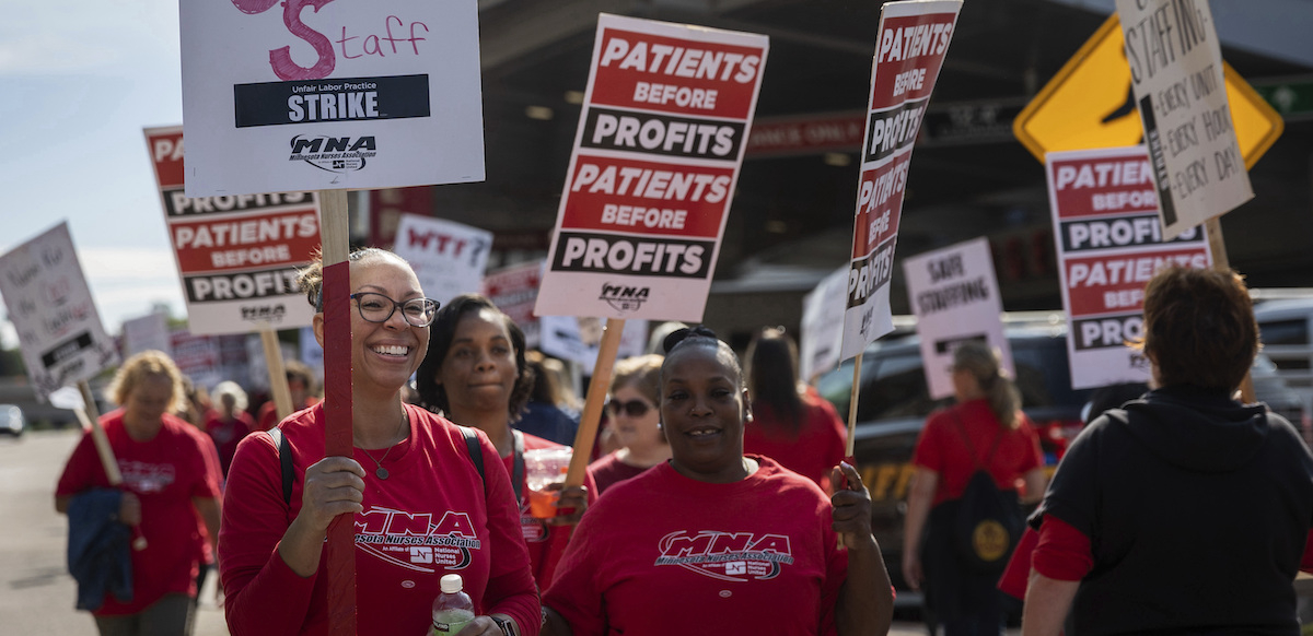 a group of smiling nurses, mostly African American, in red shirts hold signs saying ‘patients before profits’