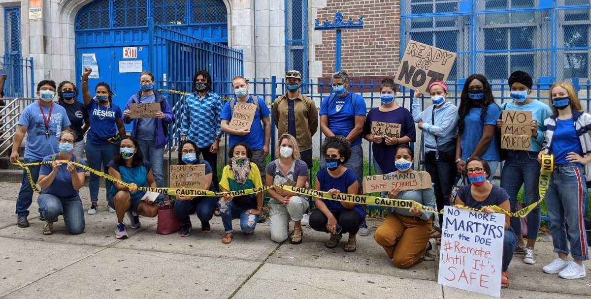 21 adults in masks outside a school. Together they hold "CAUTION" tape. Handmade signs say "100% remote start," "no mas muertes," "ready or not," "we demand safe schools," "no more martyrs for the DOE #Remote until it's safe" and "people got sick in this buidling."