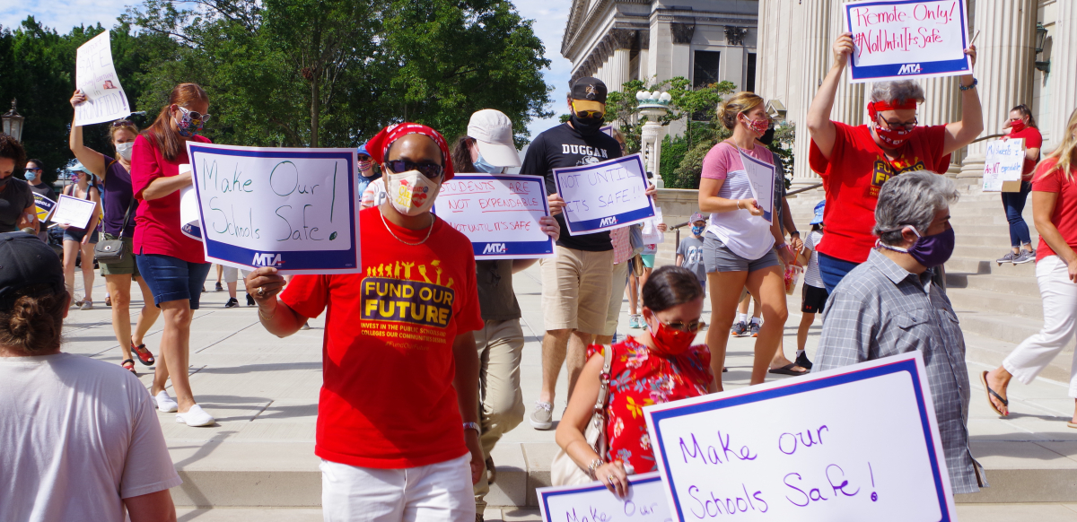 Teachers wearing masks hold signs that say Make Our Schools Safe and Not Until It's Safe.