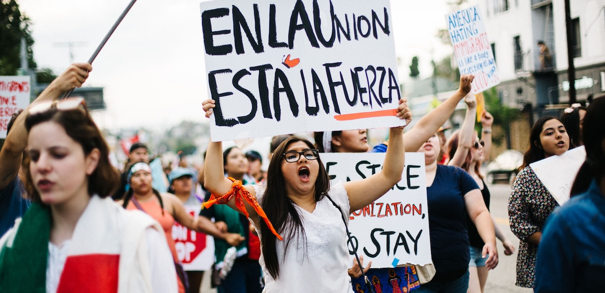 A group marches, one woman holds a sign above her head in Spanish: “En La Union Esta La Fuerza,” with a butterfly on it. Meaning “our strength lies in unity.”