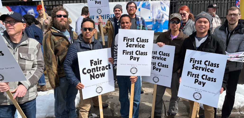 A dozen people, mostly men, face the camera resolutely on a sunny cold day outside a post office. Several are holding printed signs that say "Fair contract now" or "First class service, first class pay" with the NALC Branch 9 logo.