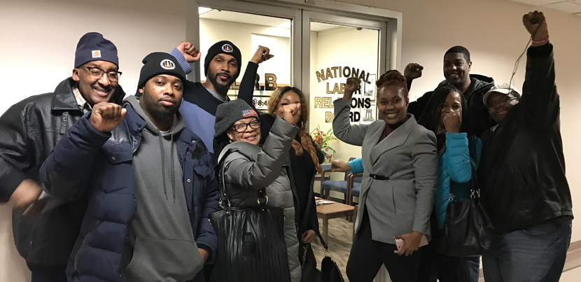 Nine Black film industry parking attendants in coats, smiling with fists up, indoors, in front of an NLRB office in New York City.