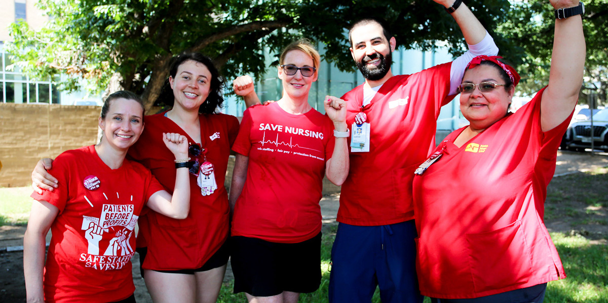 A group of five nurses in red shirts pose with their fists raised.