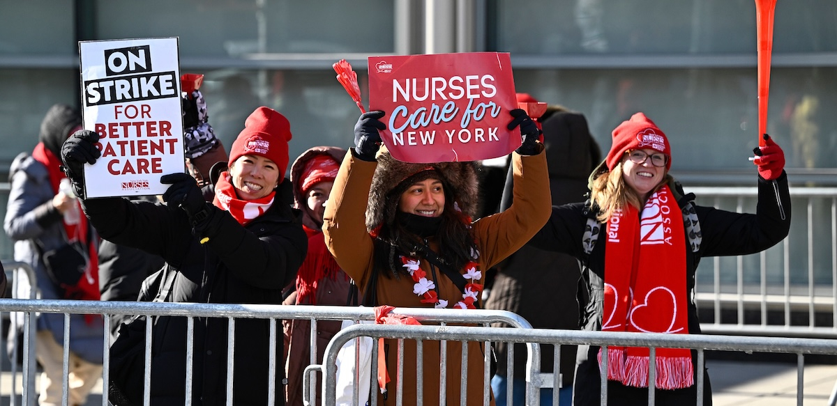 Three smiling nurses stand against a barrier with signs about patient care